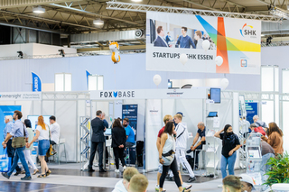 Menschen gehen in der Messehalle umher, ein Banner mit Startups SHK Essen hängt von der Decke.